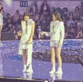 two women standing on a stage in front of a crowd with the number 112 on the wall behind them