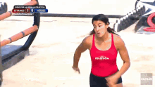 a woman in a red tank top that says exatlon is running on a beach