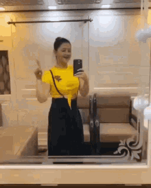 a woman is taking a selfie in front of a mirror while wearing a yellow shirt and black pants .