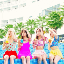 four women are sitting on the edge of a swimming pool holding drinks .