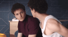 a man in a white tank top sits next to another man in a burgundy shirt