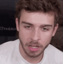 a young man with a beard is wearing a white t-shirt and looking at the camera .