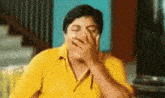 a man in a yellow shirt is laughing and covering his face with his hand .