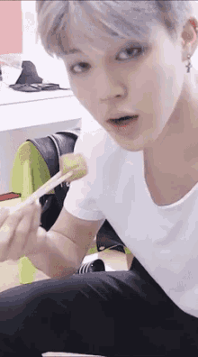 a young man in a white shirt is sitting down eating food with chopsticks .