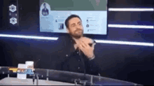 a man with a beard is sitting at a desk in front of a tv screen .
