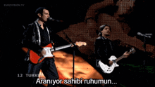 a man singing into a microphone while another man plays guitar in front of a screen that says eurovision.tv