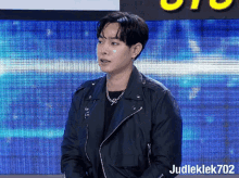 a man in a black jacket is standing in front of a large screen with judleklek702 written on it