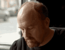 a bald man with a beard is sitting at a table in front of a window with his eyes closed .