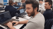 a man with a beard is typing on a laptop in a classroom