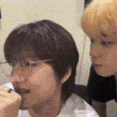 two young men are sitting next to each other in a room . one of the men has glasses on and the other has short hair .