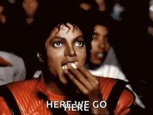 a man in a red jacket is eating popcorn while watching a movie and says `` here we go '' .