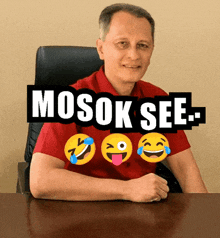 a man in a red shirt sits at a desk with a sign that says mosok see behind him