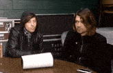 two men sit at a table in front of a sign that says taki