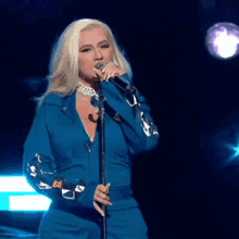 a blonde woman singing into a microphone in a blue jumpsuit
