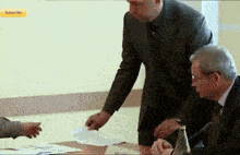 a man in a suit is pointing at a piece of paper with a subscribe button in the corner