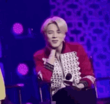 a man in a red jacket is sitting in front of a microphone on a stage in front of a purple background .