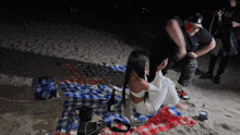 a woman sits on a plaid blanket on the beach with a man standing behind her