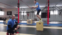 a man is jumping over a box in a gym while a man watches .