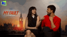 a man and woman are sitting next to each other in front of a poster for my fault london