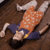 a man wearing a purple shirt and orange apron is laying on the floor with his eyes closed