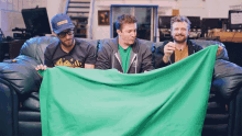 three men are sitting on a couch holding a green cloth and one of them is wearing a blue hat with the word columbia on it