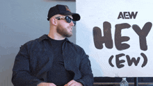 a man wearing sunglasses and a hat stands in front of a sign that says hey ( ew )
