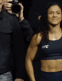 a woman wearing a blue ufc top stands in front of a man holding a camera