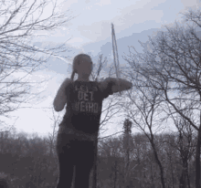 a woman in a black shirt that says let 's get weird is standing in the woods holding a sword .