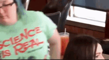 a woman wearing a green shirt that says science is real is sitting at a table with a drink .
