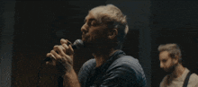 a man with a beard is singing into a microphone while another man stands behind him playing a guitar .