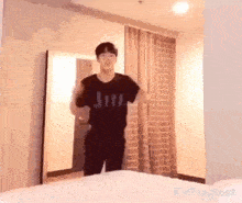 a young man in a black shirt is standing in a room next to a bed .