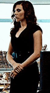 a woman in a black dress stands with her hands folded in front of a window