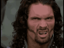 a man with long hair and a beard is making a funny face and the words alright are visible in the corner .