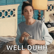 a woman is sitting on a bed holding a glass of water and saying well duh .