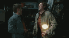 two men are standing next to each other and one has a light in his hand and the other has a cw logo on his shirt