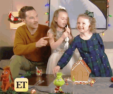 a man and two little girls are sitting at a table with a gingerbread house in front of them and an et logo