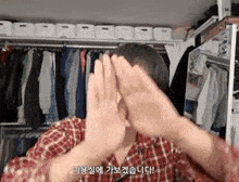 a man in a plaid shirt is covering his face with his hands in front of a closet filled with clothes .