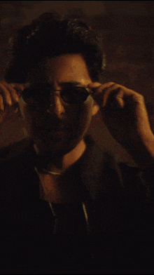 a man wearing sunglasses and a necklace stands in front of a brick wall