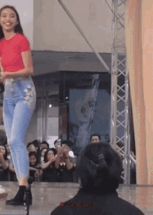 a woman in a red shirt and blue jeans stands on a stage in front of a crowd