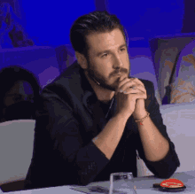 a man with a beard sits at a table with his hands folded in front of his face