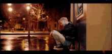 a young man is squatting down on the sidewalk in front of a building at night .