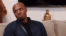 a bald man is sitting on a couch with a woman and a buddha statue in the background .