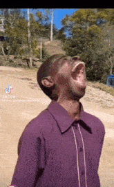 a man in a purple shirt is laughing with his mouth wide open while standing on a dirt road .