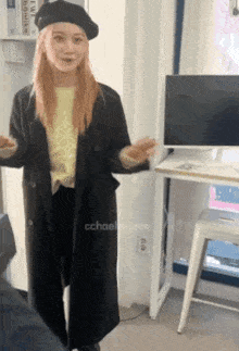 a woman wearing a black coat and a black beret stands in front of a computer monitor