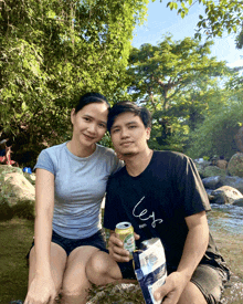 a man and a woman are sitting on a rock by a river holding a can of beer
