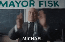 a bald man in a suit and tie is standing in front of a mayor fisk sign .