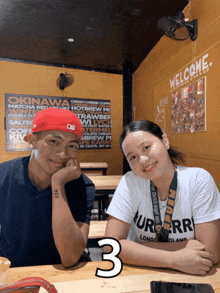 a man and a woman are sitting at a table with a welcome sign on the wall behind them