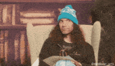 a man with long curly hair wearing a blue hat is sitting in a chair holding a piece of paper