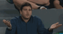 a man in a blue shirt is sitting in a stadium and making a funny face with his hand .