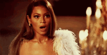 a woman in a feathered dress is standing in front of a chandelier and making a surprised face .